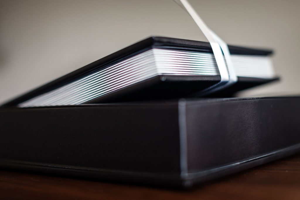 A close-up of a black photo album with thick pages, partially lifted from its matching black case by a white ribbon, resting on a wooden surface.