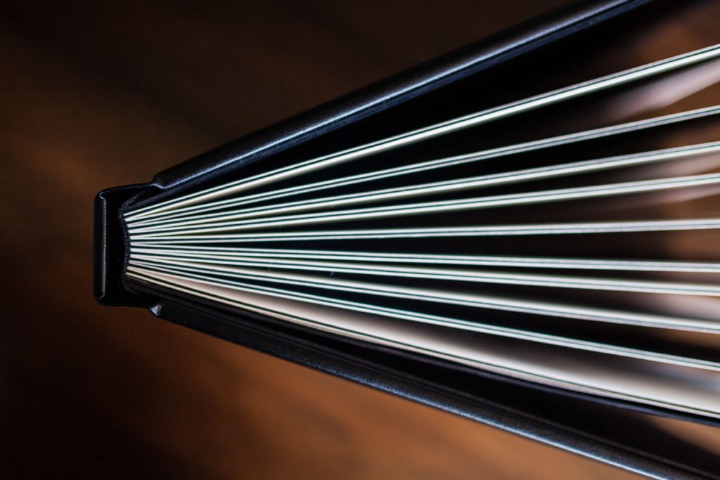 Close-up view of an open book, photographed from above at an angle, showing the edges of the pages fanned out and the black cover along the spine against a blurred, dark background.