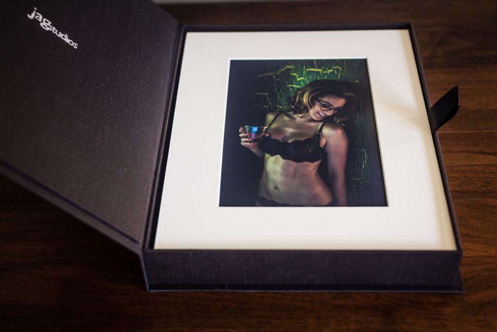 A photo print in a black presentation box shows a woman in black lingerie holding a drink and smiling, with green lighting in the background. The box rests on a wooden surface.