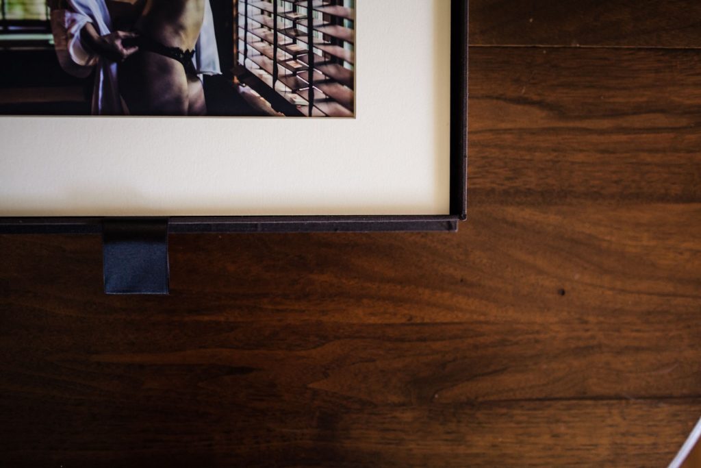 A close-up of a framed photograph on a wooden surface, showing part of an image with a person standing near window blinds, with the rest of the frame and surface visible.