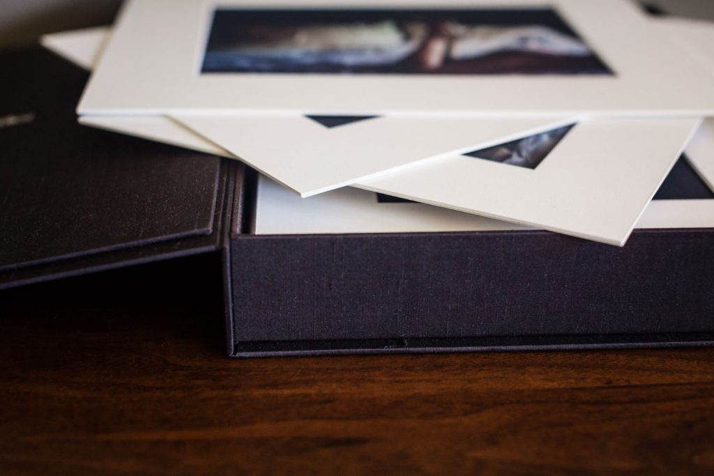 A close-up of a black box containing several matted photo prints, with some mats stacked on top and edges visible, resting on a wooden surface.