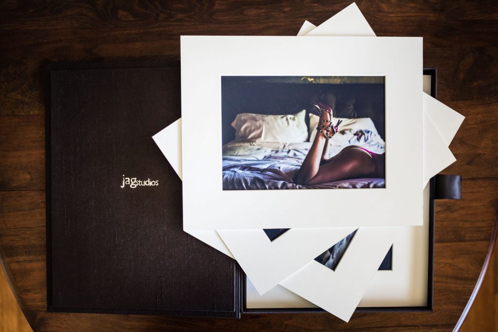 A photo print of a woman lying on a bed with her legs raised, placed atop white photo mats and a black box labeled jagstudios on a wooden surface.