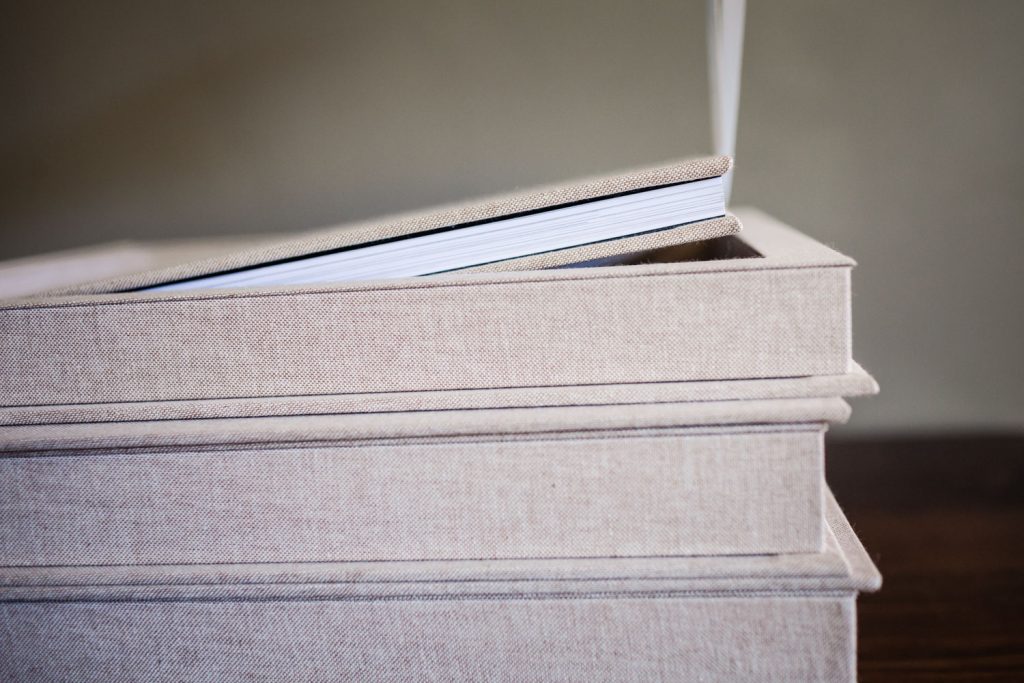 A close-up of three beige fabric-covered photo albums stacked on a dark wooden surface, with the top album slightly open and its pages visible.