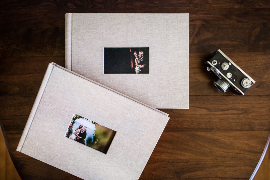 Two beige photo albums with small photos on their covers rest on a wooden table next to a vintage black and silver camera.