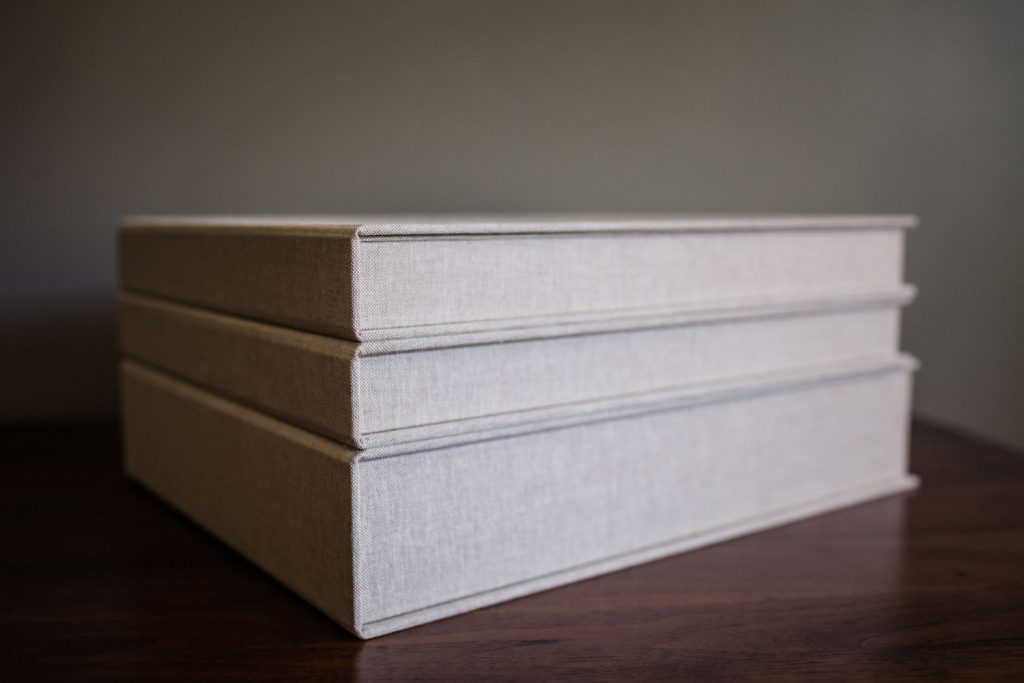 A stack of three beige, fabric-covered photo albums or boxes sits on a wooden surface against a plain, neutral background.