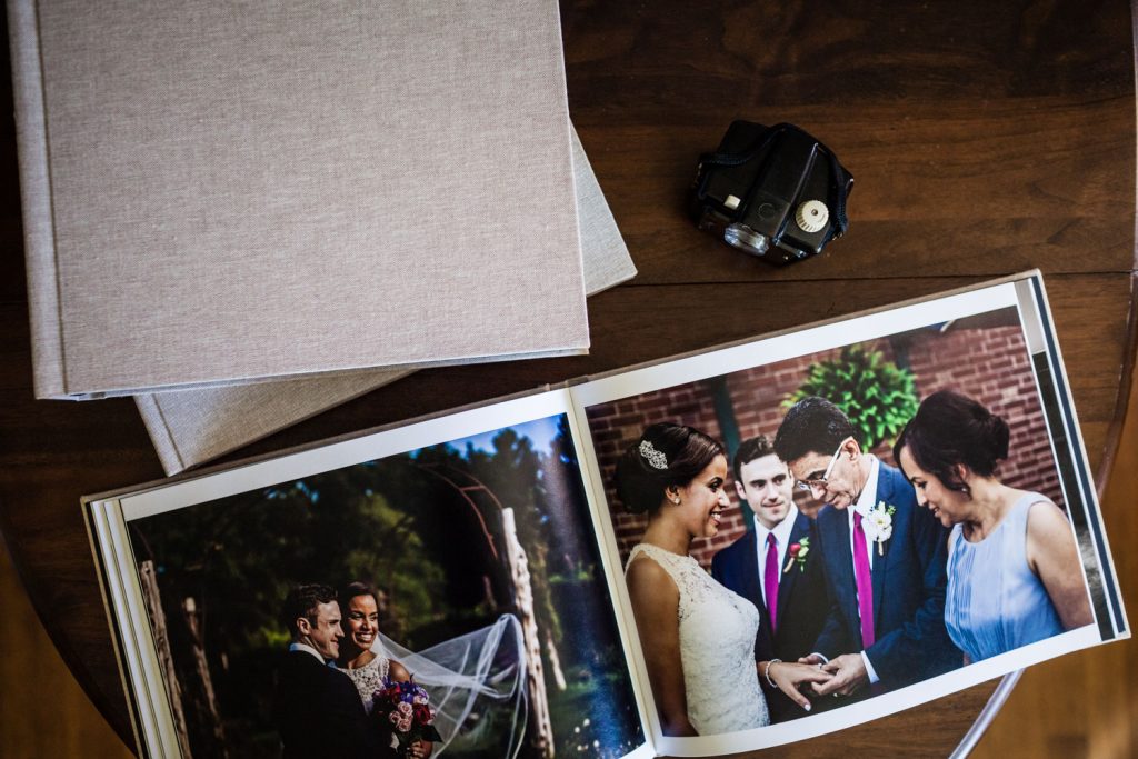 An open wedding photo album sits on a wooden table, showing joyful moments from the ceremony. Nearby are two closed albums and a camera, capturing a sense of celebration and memories.