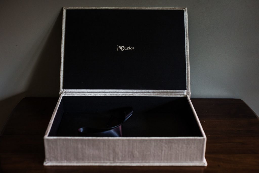 A beige textured box with a black interior sits open on a dark wooden surface. Inside the lid, jagstudios is printed in white. A black object rests at the bottom of the box.