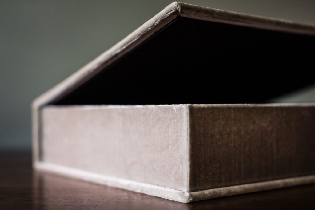 A close-up view of a partially open, rectangular, velvet-covered box resting on a wooden surface, with the lid propped up at an angle.