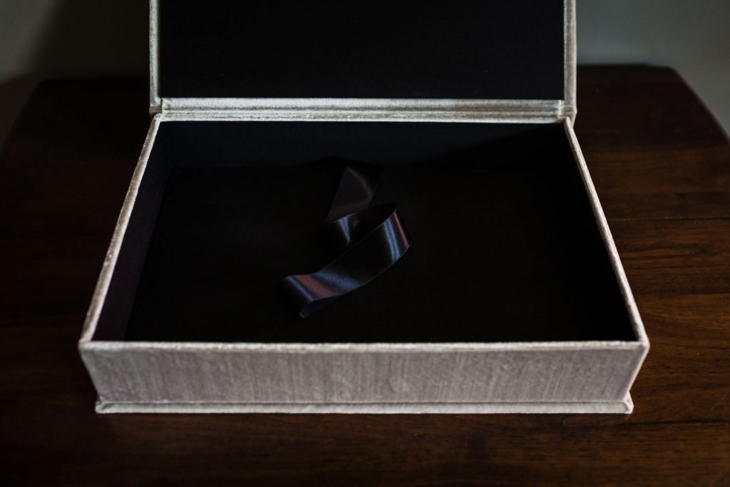 An open, empty beige velvet box with a black interior and a single dark ribbon lying inside, placed on a wooden surface.
