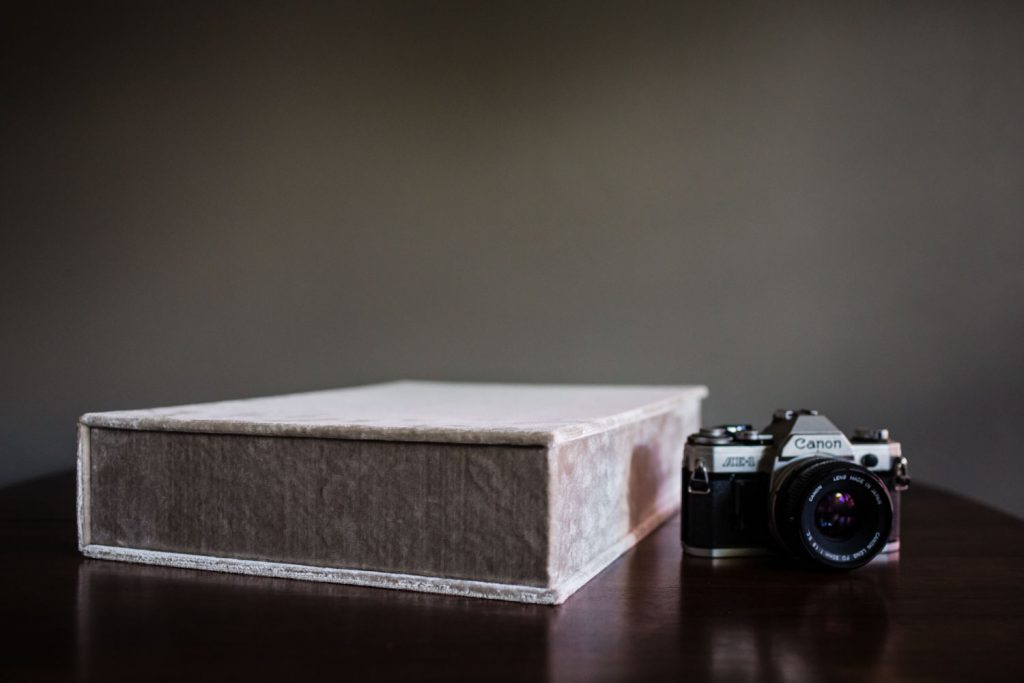 A vintage Canon film camera sits on a dark wooden surface next to a large, closed, light gray box, against a plain, dark background.