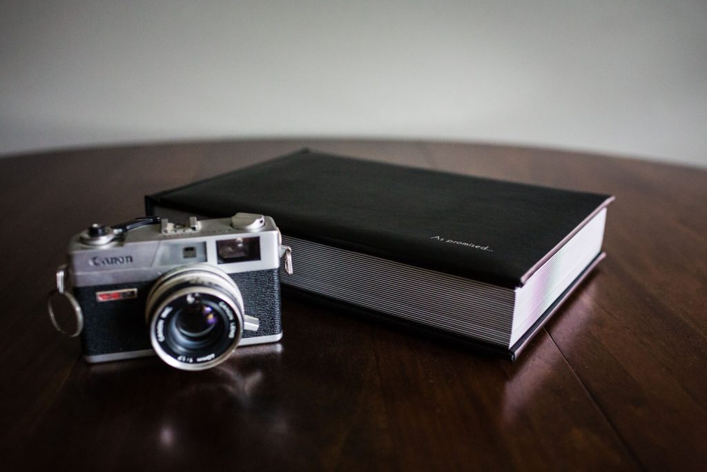 A vintage Canon film camera sits on a wooden table next to a closed, thick black photo album with “A portrait.” written on the cover.