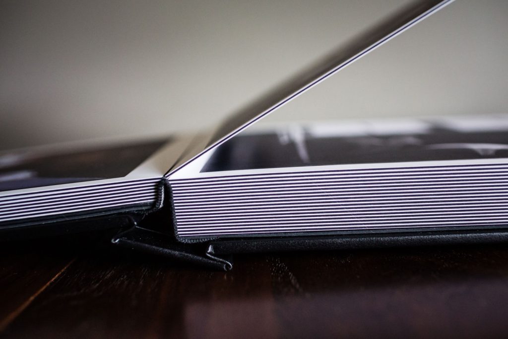 A close-up side view of an open photo album resting on a wooden surface, showing thick, white-edged pages with images printed inside.