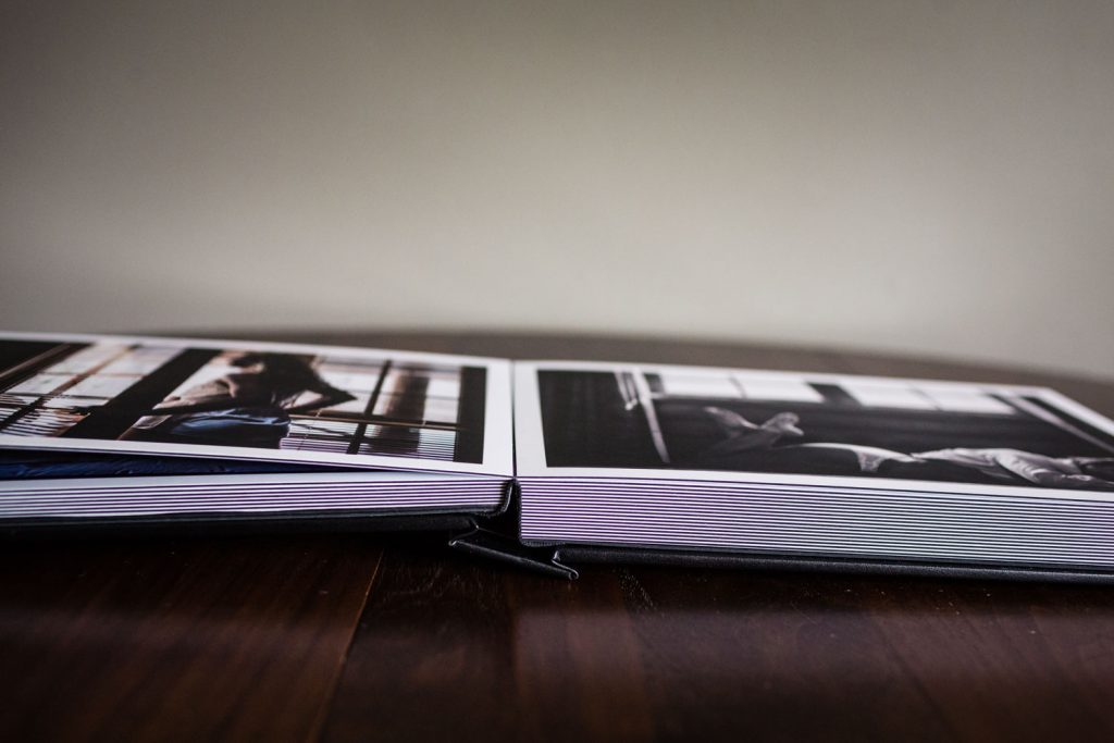 A photo album opened on a wooden table, displaying two pages with black and white photographs of people sitting near windows, with a neutral background behind the album.