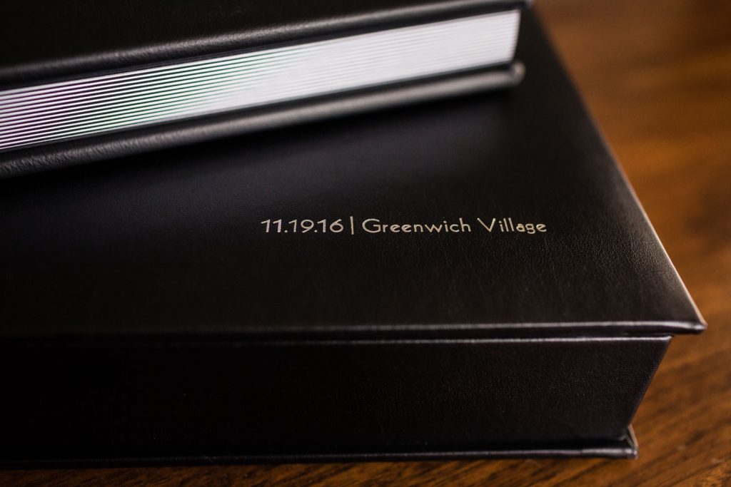 A close-up of a black leather-bound book with 11.19.16 | Greenwich Village embossed in gold on the cover, resting on a wooden surface.