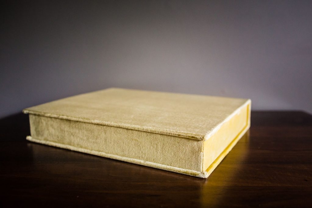 A closed, light beige book with a fabric cover sits on a dark wooden surface against a plain gray background.
