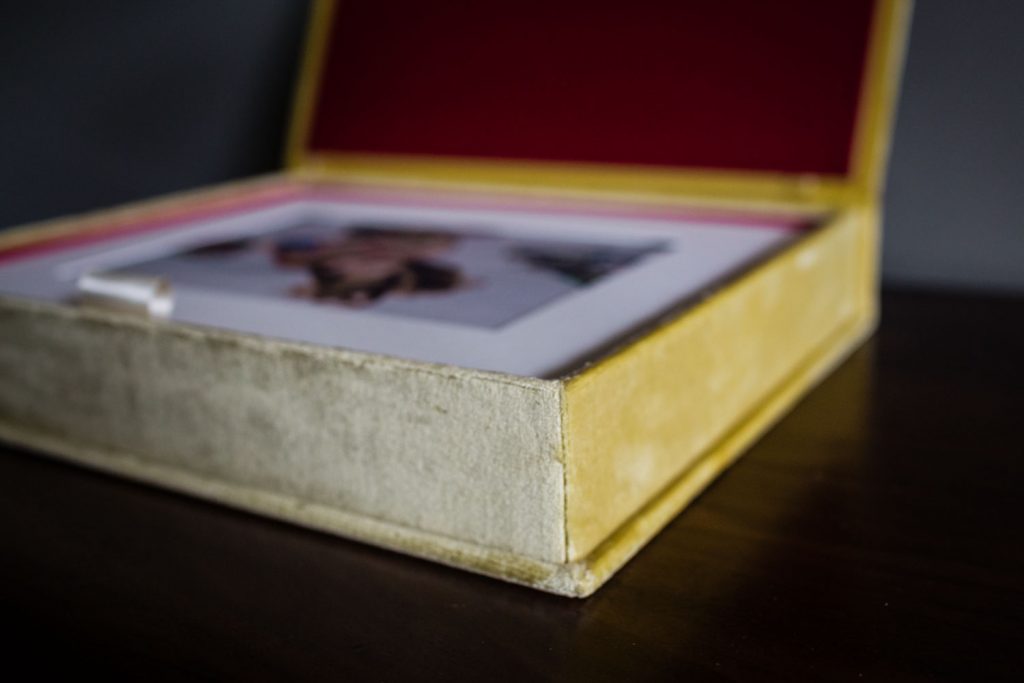 A close-up of a yellow velvet photo album box with a red interior, resting on a dark wooden surface. The box is open, revealing a photograph inside, but the image is out of focus.