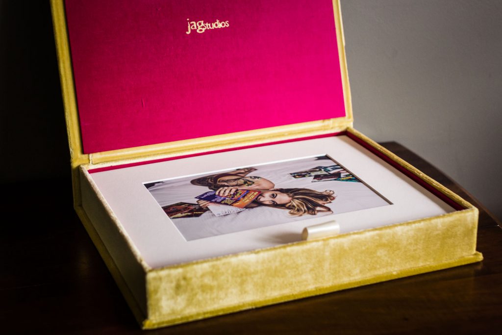 A yellow velvet box with a pink interior, labeled jagstudios, holds a matted photo of a woman lying down, partially covered by a colorful patterned blanket. The box rests on a wooden surface.