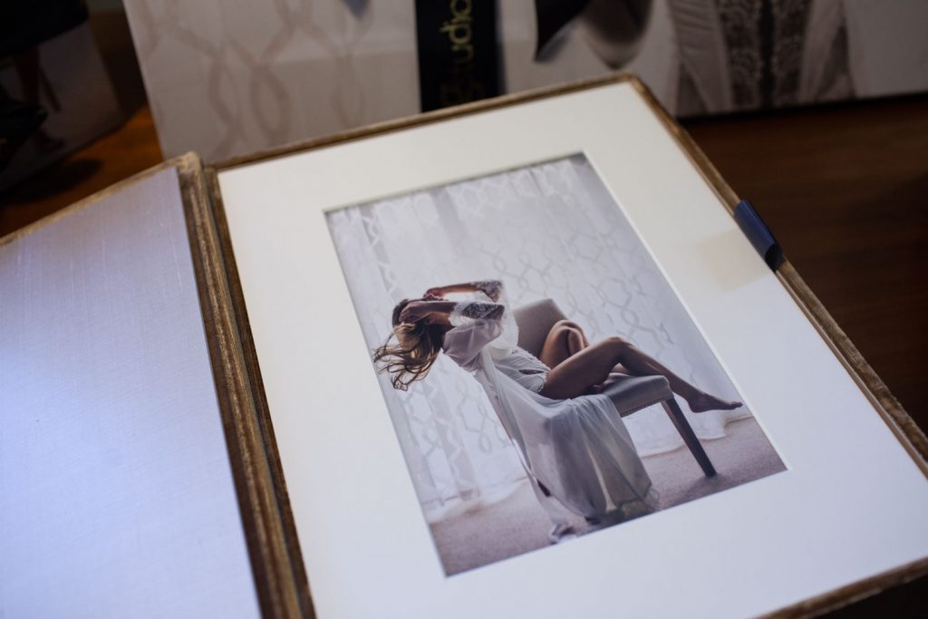 A photo album open to a framed image of a woman in a sheer robe posing on a chair near a window with sheer curtains, her hair flowing and one arm raised to her head.