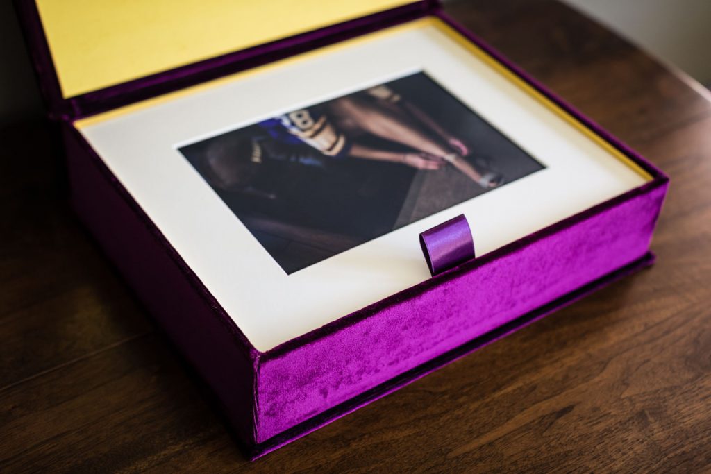 A vibrant purple velvet photo box with a yellow interior sits open on a wooden table. Inside, a matted photograph of a person’s legs and running shoes is displayed.
