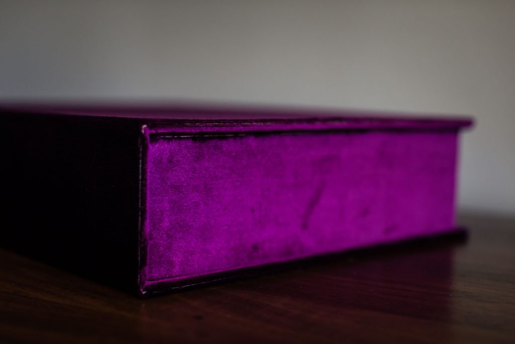 A velvet-covered, purple box sits on a wooden surface, photographed from the side against a plain, blurred background.