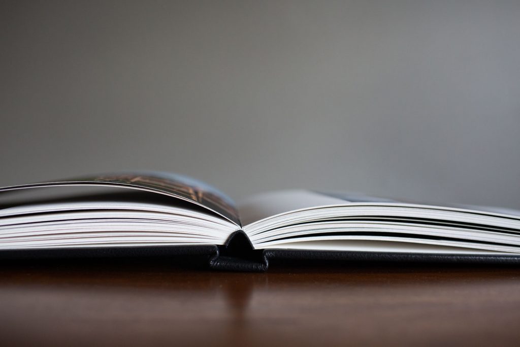 An open book lies flat on a wooden surface, with its pages slightly curved upward and a blurred gray background.