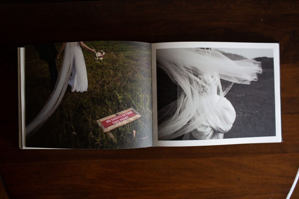 An open photo album on a wooden table shows two wedding photos: one in color with a bride holding a bouquet, and one in black and white with her flowing veil. A sign reads, “No guest book, just photos.”.