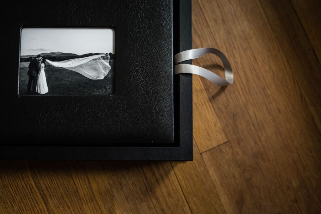A black photo album with a silver ribbon lies on a wooden surface. The cover features a small black-and-white photo of a bride and groom outdoors, with the brides veil blowing in the wind.