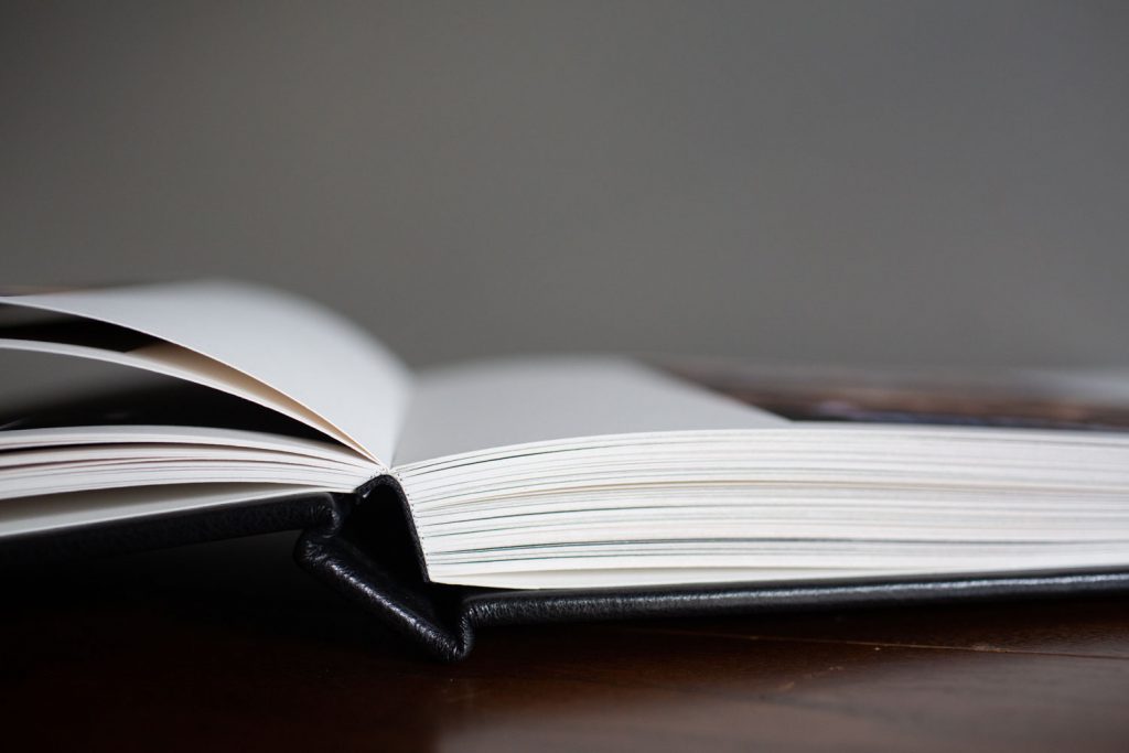 A close-up view of an open hardcover book lying on a dark wooden surface, with pages slightly fanned and a blurred gray background.