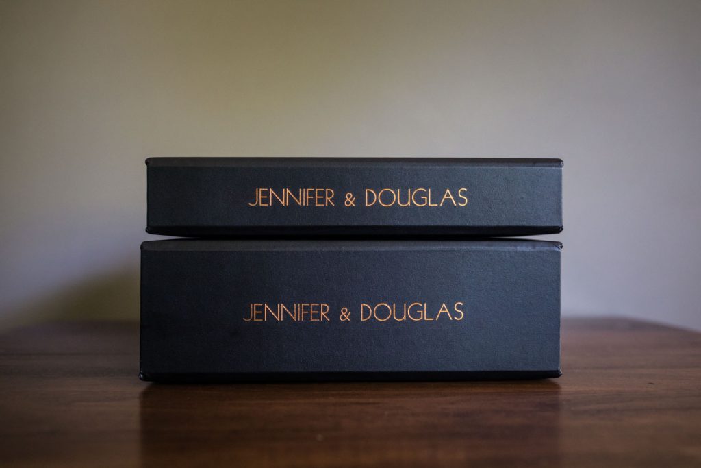 Two black boxes stacked on a wooden surface, each with the names Jennifer & Douglas written in gold lettering on the side. The background is plain and neutral.