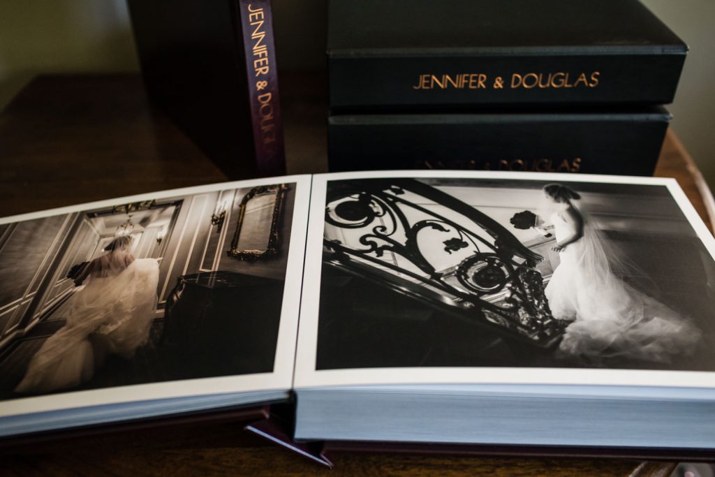 An open photo album displays black-and-white wedding photos of a bride on a staircase. Next to it are closed albums labeled JENNIFER & DOUGLAS in gold letters. All albums are on a wooden surface.