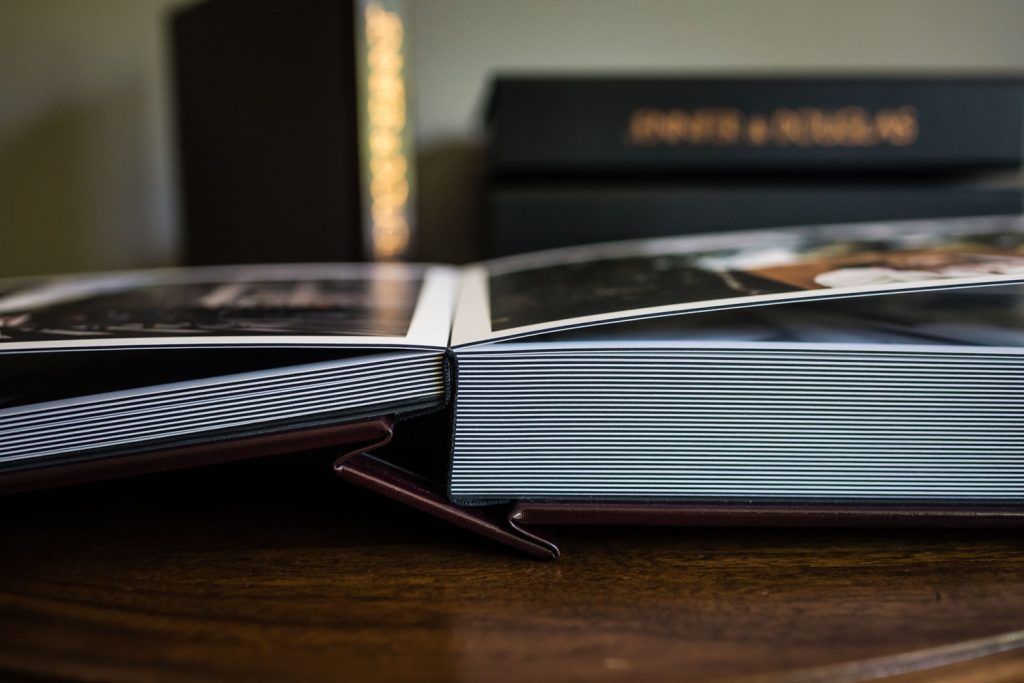 A close-up view of an open photo album resting on a wooden surface, with its thick, glossy pages visible. Blurred books or albums are stacked in the background.