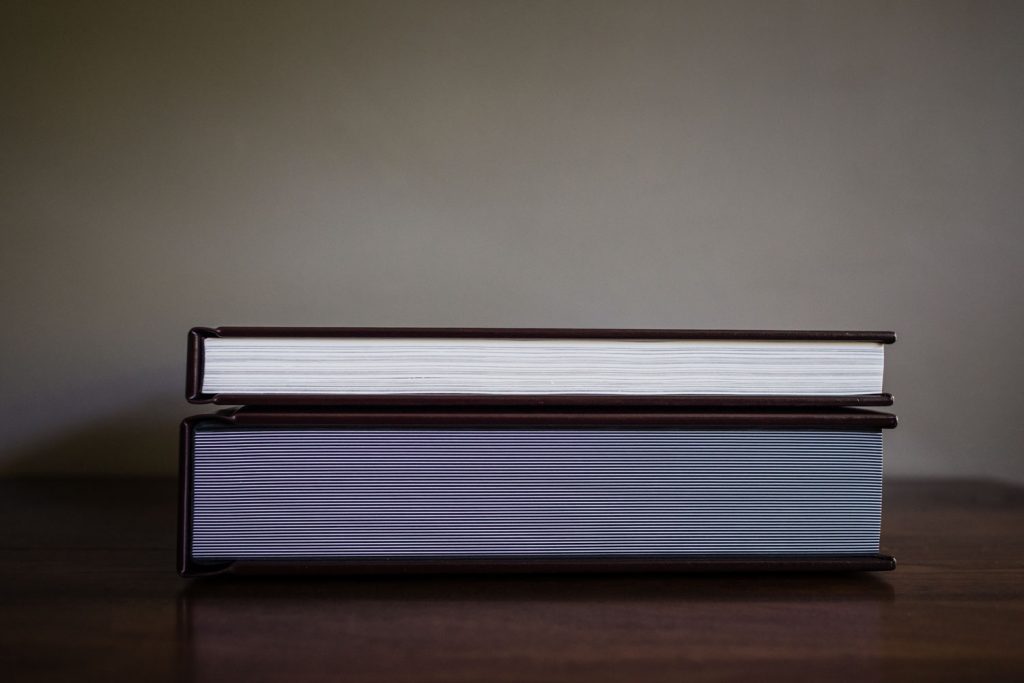Two closed hardcover books stacked on top of each other on a wooden surface, viewed from the side, with a plain, neutral-colored background.