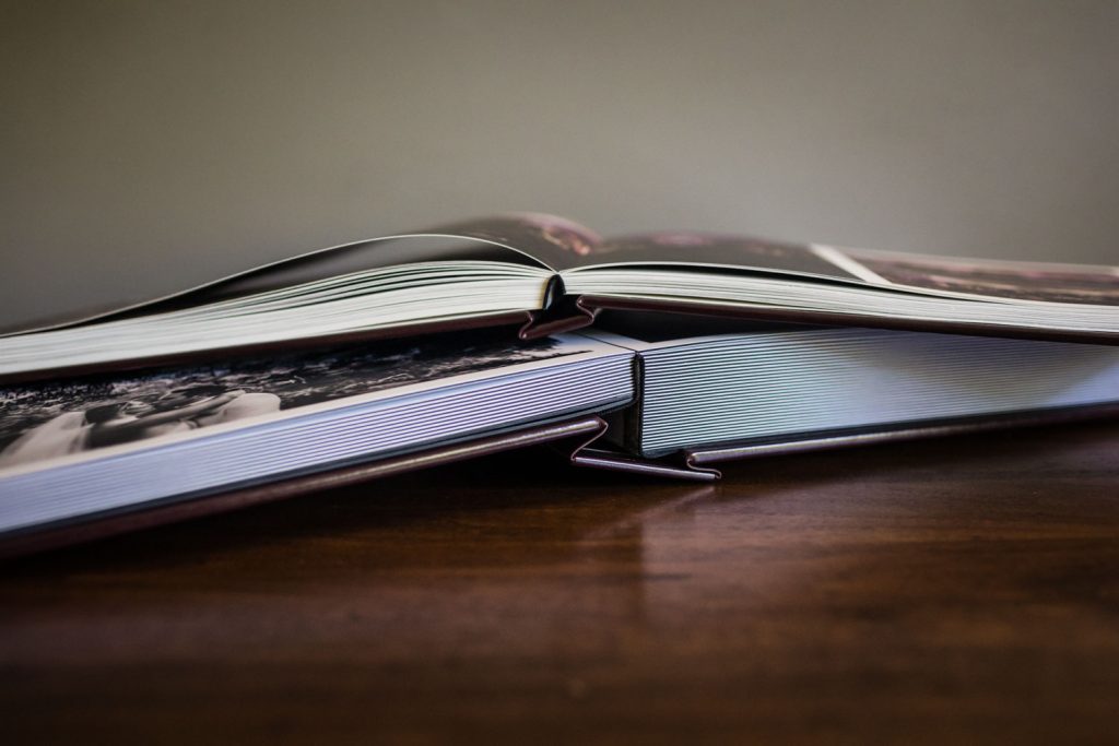 Close-up of two open photo albums lying on a wooden surface, with pages slightly curved and overlapping each other, showing glimpses of black and white photographs inside.