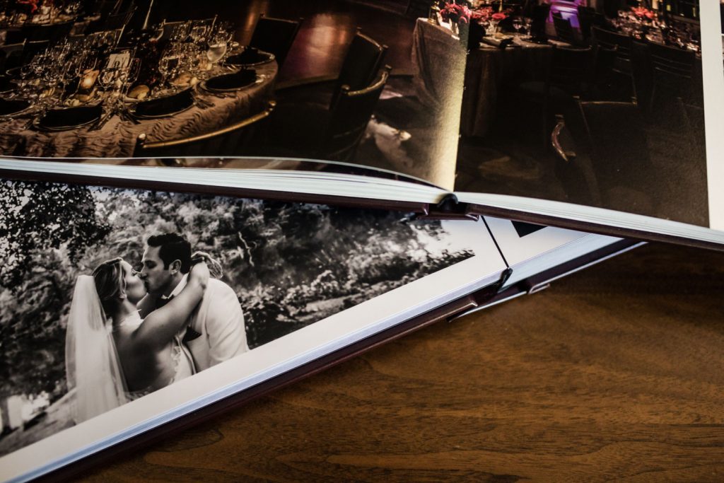An open wedding photo album on a wooden table, displaying a black-and-white photo of a bride and groom kissing and a color photo of a decorated reception table.