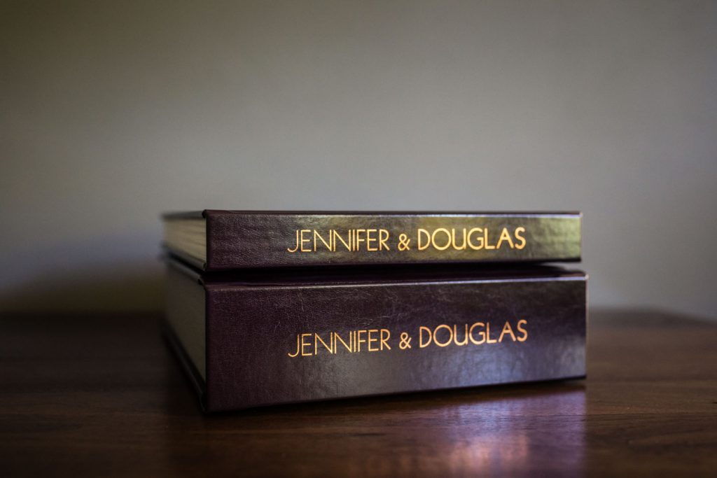 Two dark brown hardcover photo albums stacked on a wooden surface, both with JENNIFER & DOUGLAS written in gold lettering on the spines. The background is softly blurred.
