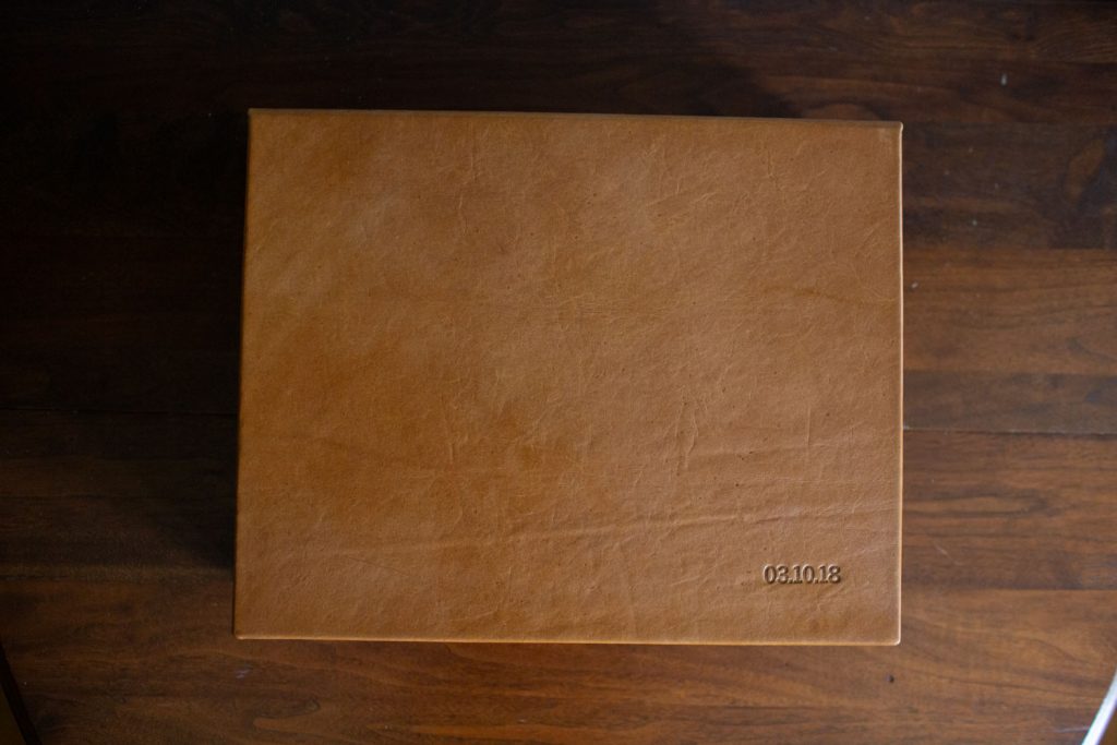 A closed, light brown leather-bound book or album sits on a dark wooden surface. The date 03.10.18 is embossed in the lower right corner of the cover.