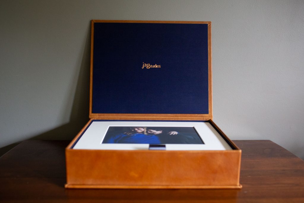 A brown leather photo box with a navy blue interior, displaying a framed photograph of two people inside, is open on a wooden table against a gray wall. The box lid has jagstudios written in gold text.