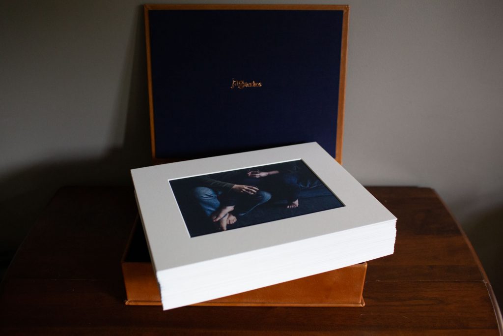 A stack of photo prints with a white border sits on top of an open brown box with a dark blue interior, displayed on a wooden table. The top print shows a close-up of two people sitting together.