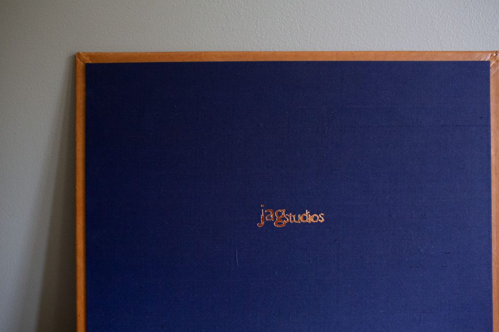 A close-up of a navy blue textured board with a brown wooden frame and the text ja studios embroidered in orange at the center. The board is leaning against a light gray wall.