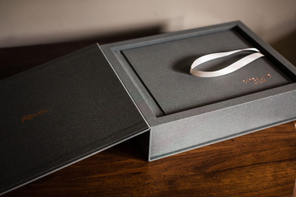A gray, fabric-covered box with gold embossed writing sits open on a wooden surface; inside, a matching folder with a white ribbon is neatly placed.