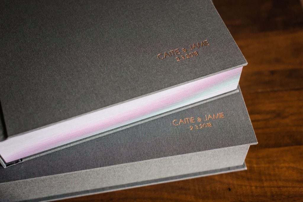 Two gray hardcover books with gold embossed text on the covers. The top book reads CATIE & JAMIE 2.5.2018, and the bottom book reads CATIE & JAMIE 9.3.2016. The books are on a wooden surface.