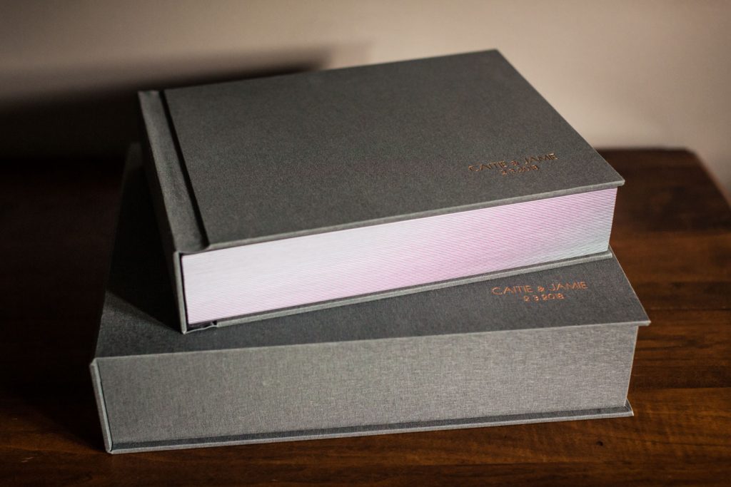 Two large gray photo albums with gold embossed text on the covers are stacked on a wooden table. The top albums pages are slightly open, revealing a glimpse of the inside.