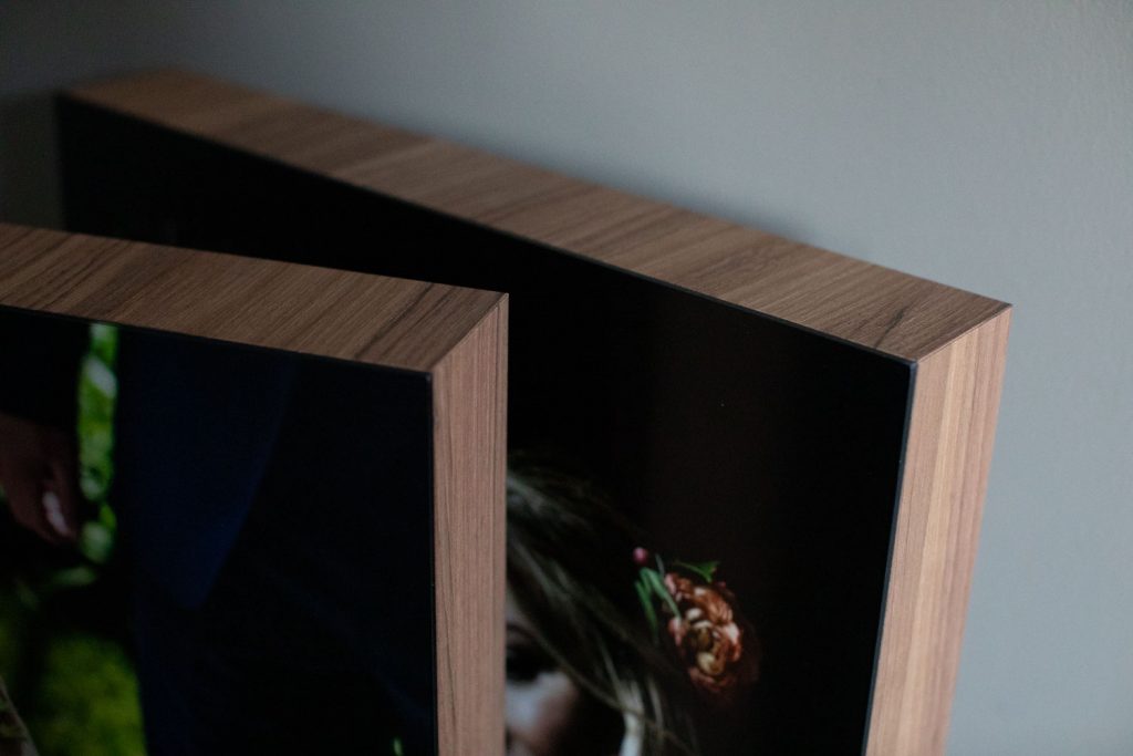 Two wood-framed photo prints leaning against a wall; one shows part of a person’s face and a flower, while the other is mostly obscured. The photo focuses on the angled wood edges and frame detail.