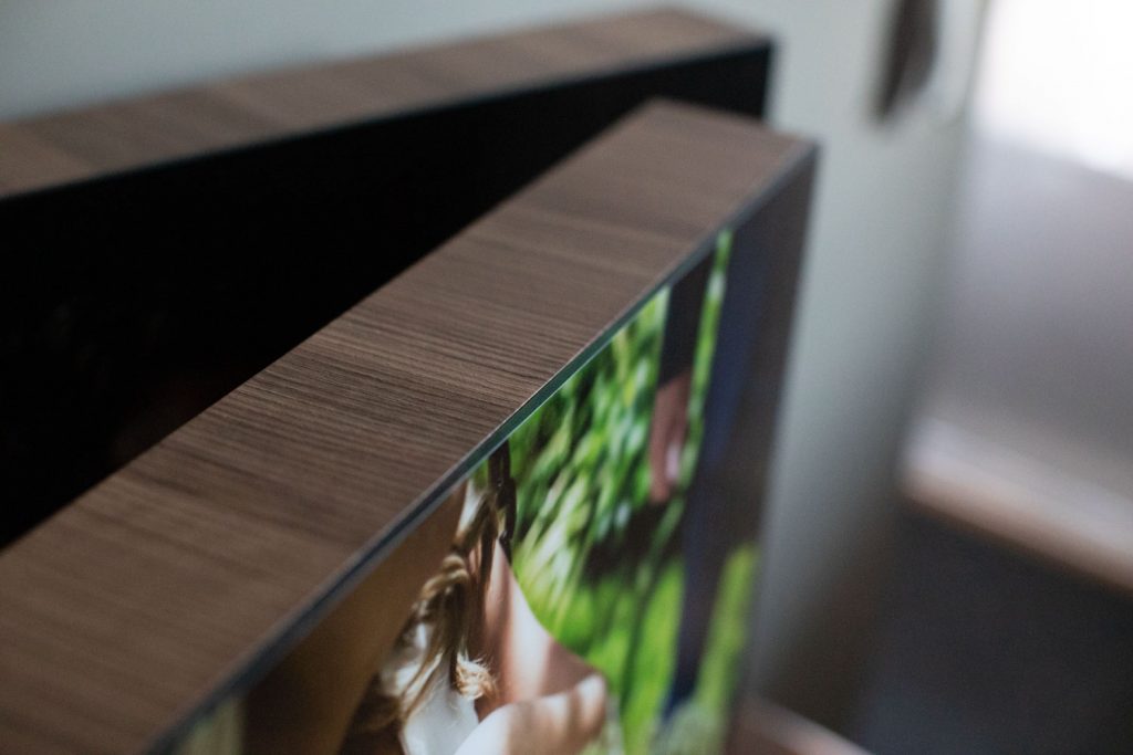 Close-up view of the top edge of a flat-screen TV with a wood-textured frame, displaying a blurred image that includes greenery and part of a person. The background is softly out of focus.