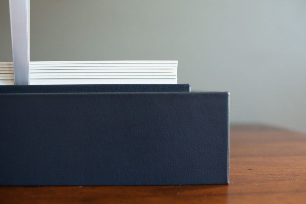 A close-up of the edge of a dark blue hardcover book with a white ribbon bookmark, resting on a wooden surface against a plain gray background.