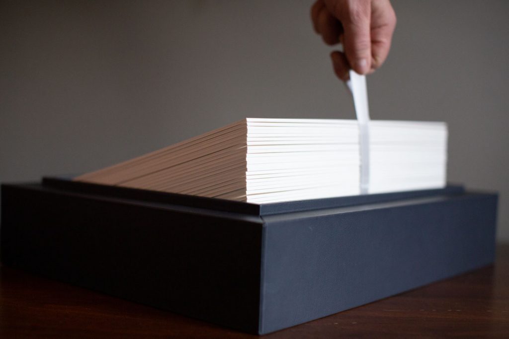 A hand lifts a strip of paper from a neatly stacked pile of white index cards inside a black box on a wooden surface.