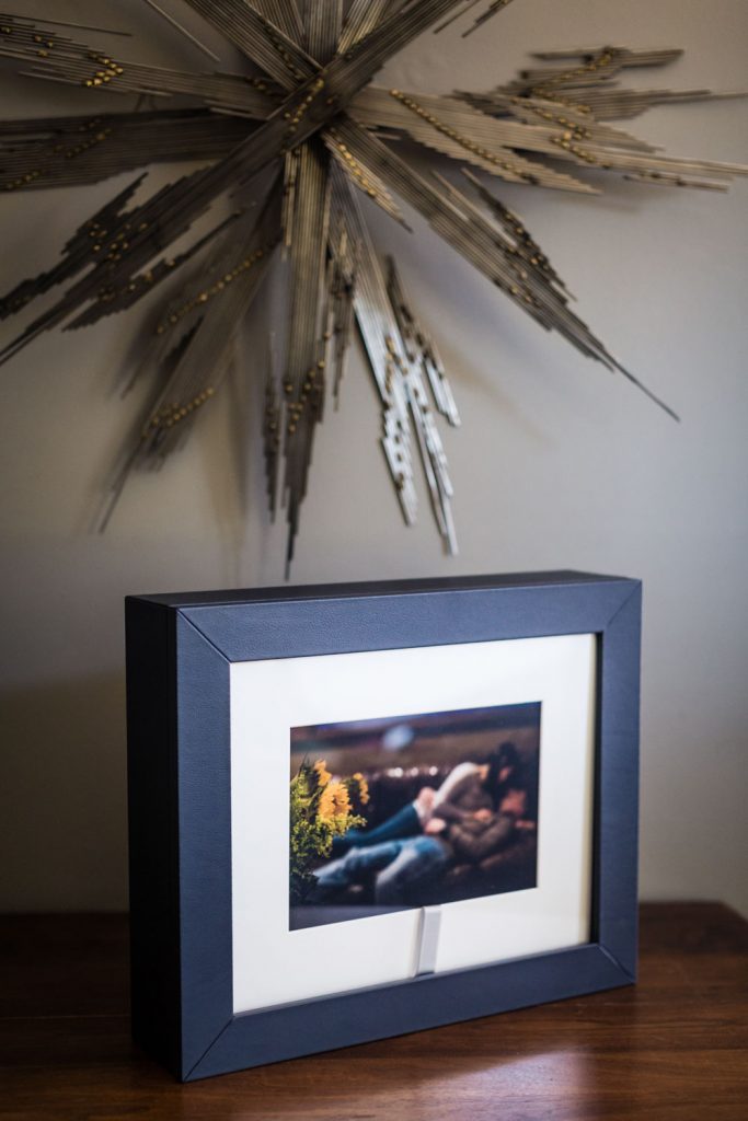 A framed photo of two people sitting on a couch is displayed on a wooden surface. Behind it, a large, decorative metallic starburst sculpture is mounted on a neutral-colored wall.