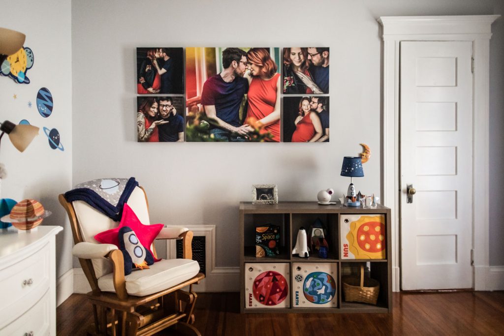 A cozy nursery with a rocking chair, a star pillow, shelves with toys and books, space-themed decorations, and family photos on the wall above the shelves. A closed white door is on the right side of the room.