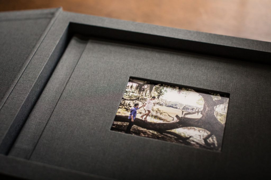 A gray photo album lies open, displaying a single photo of two children playing on a large, fallen tree branch in a sunlit outdoor setting.