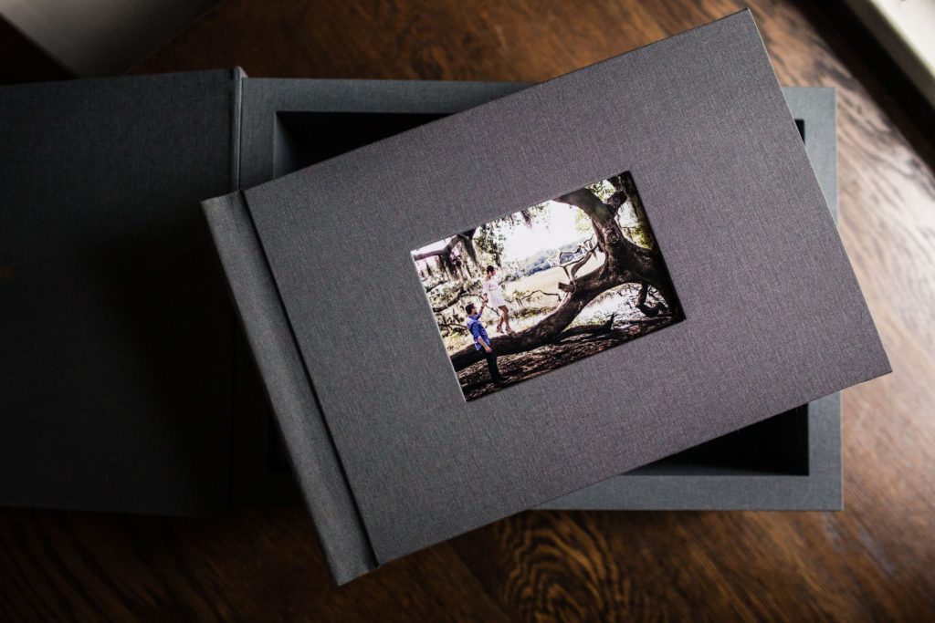 A gray photo album with a small cover photo of a person standing near a large tree is resting on top of an open box on a wooden surface. Light shines in from a nearby window.
